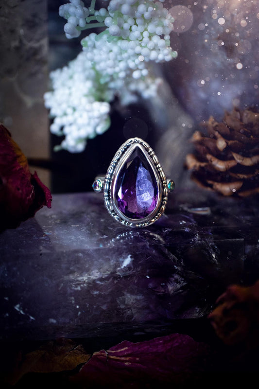 Handmade statement ring crafted from sterling silver in a striking gothic design. This bold piece features a stunning faceted amethyst as the central gemstone, radiating rich purple hues. Flanking the amethyst are sparkling aurora cubic zirconia, that catch the light with a dazzling iridescence. The intricate detailing and dark romantic style give the ring a mystical feel, perfect for lovers of gothic, or alternative jewelry. Unique and eye-catching, this ring makes a powerful, meaningful gift.
