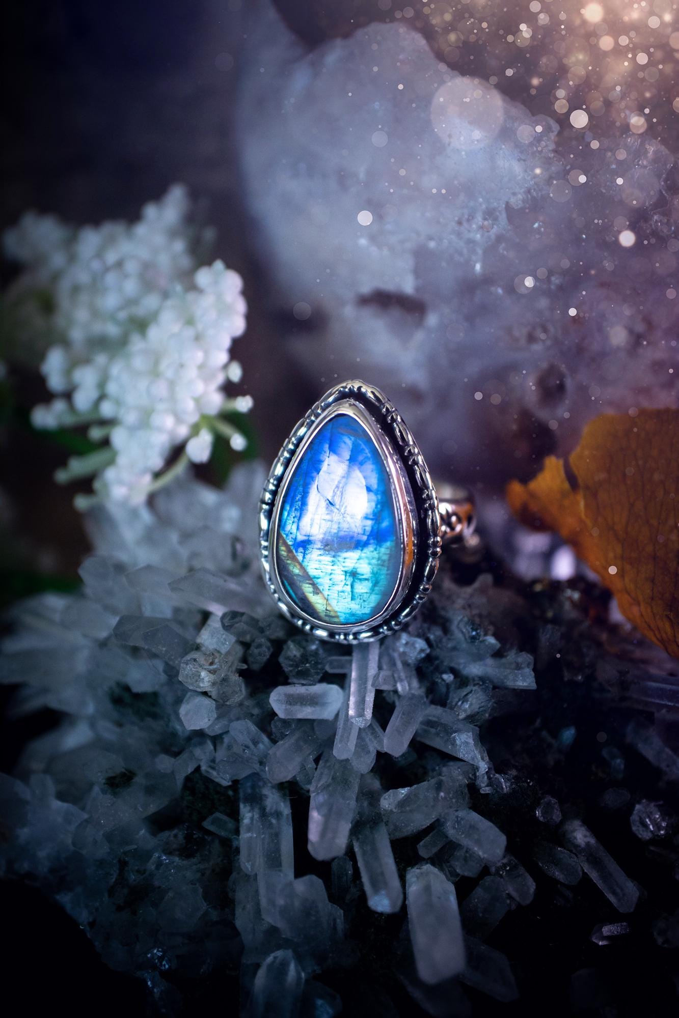 A handcrafted rainbow moonstone statement ring with a large, iridescent gemstone glowing with blue and purple flashes. The design blends gothic and whimsical "hoppy" vibes, with bespoke stamping etched into the sterling and fine silver shank. The silver band features intricate detailing, adding texture and character, while the moonstone is bezel-set, enhancing its mystical, ethereal aura. A bold and unique artisan piece with personalized charm. Ideal for crystal and jewellery lovers, h