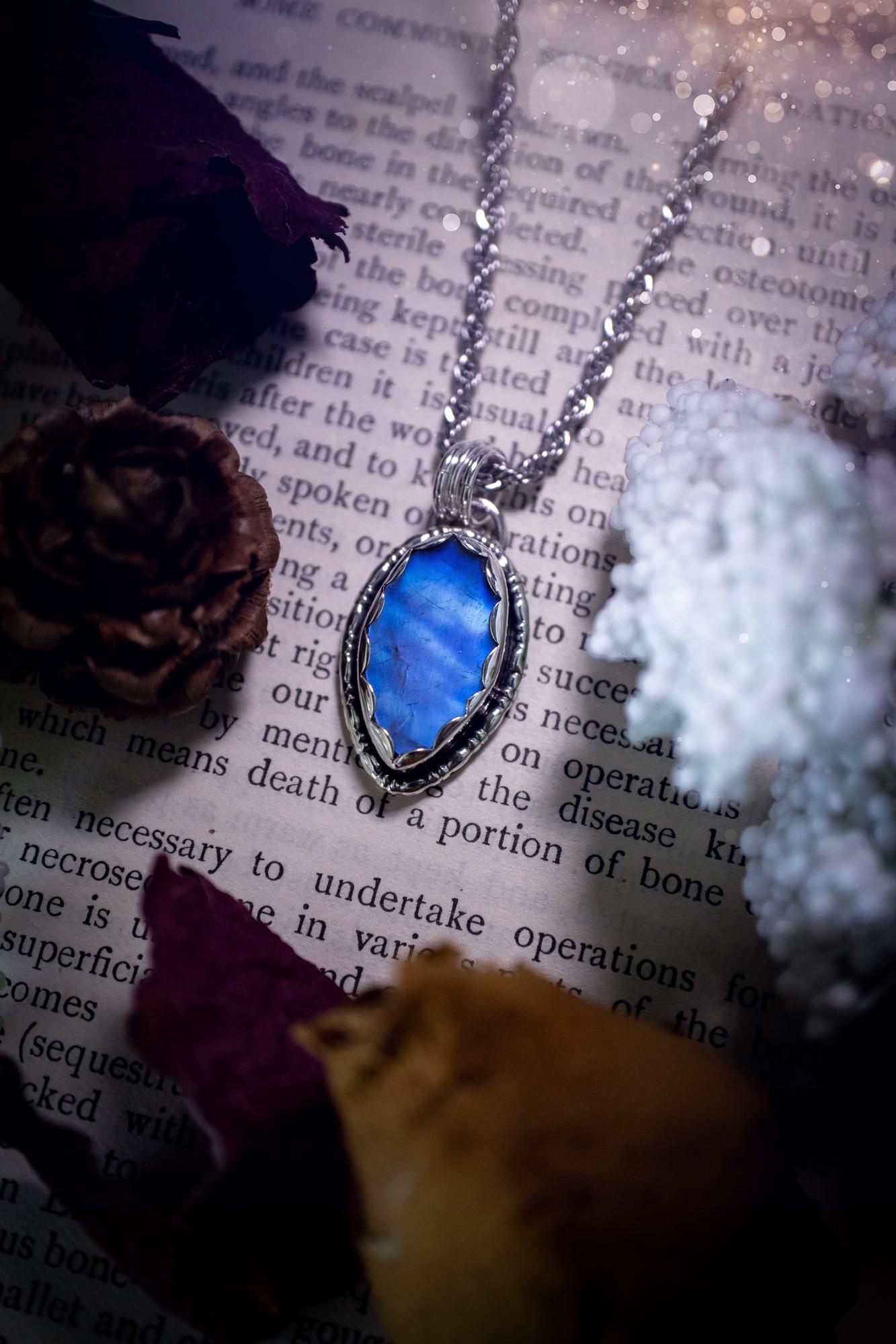 A handmade rainbow moonstone pendant necklace featuring a glowing, iridescent moonstone with blue and purple flashes. The stone is set in a scallop bezel, framed by bespoke gothic gallery wire that adds both hippy and witchy vibes. Crafted from sterling and fine silver, the pendant hangs on a silver chain, exuding mystical charm and alternative style. A one-of-a-kind piece with gothic, and magical energy. The ideal gift for crystal and jewellery lovers. Moonstone is the birthstone for June.