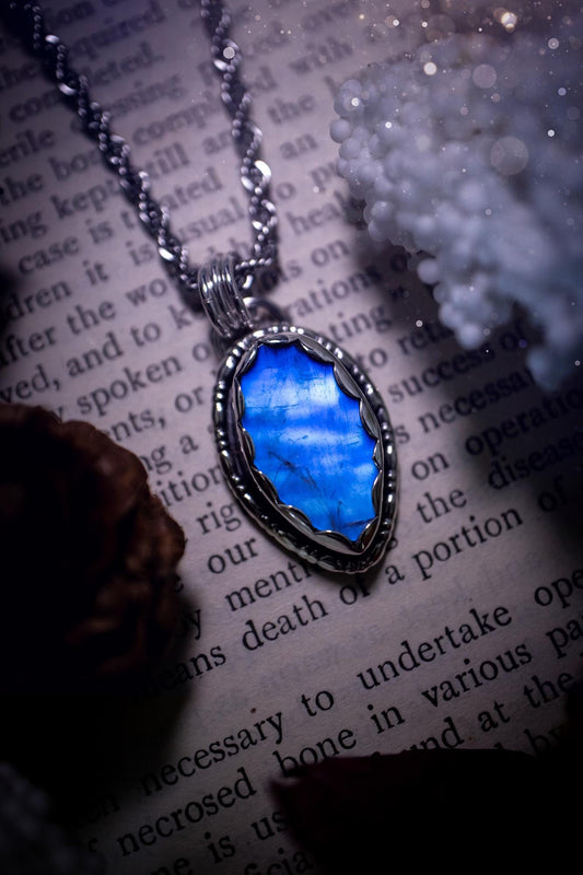 A handmade rainbow moonstone pendant necklace featuring a glowing, iridescent moonstone with blue and purple flashes. The stone is set in a scallop bezel, framed by bespoke gothic gallery wire that adds both hippy and witchy vibes. Crafted from sterling and fine silver, the pendant hangs on a silver chain, exuding mystical charm and alternative style. A one-of-a-kind piece with gothic, and magical energy. The ideal gift for crystal and jewellery lovers. Moonstone is the birthstone for June.