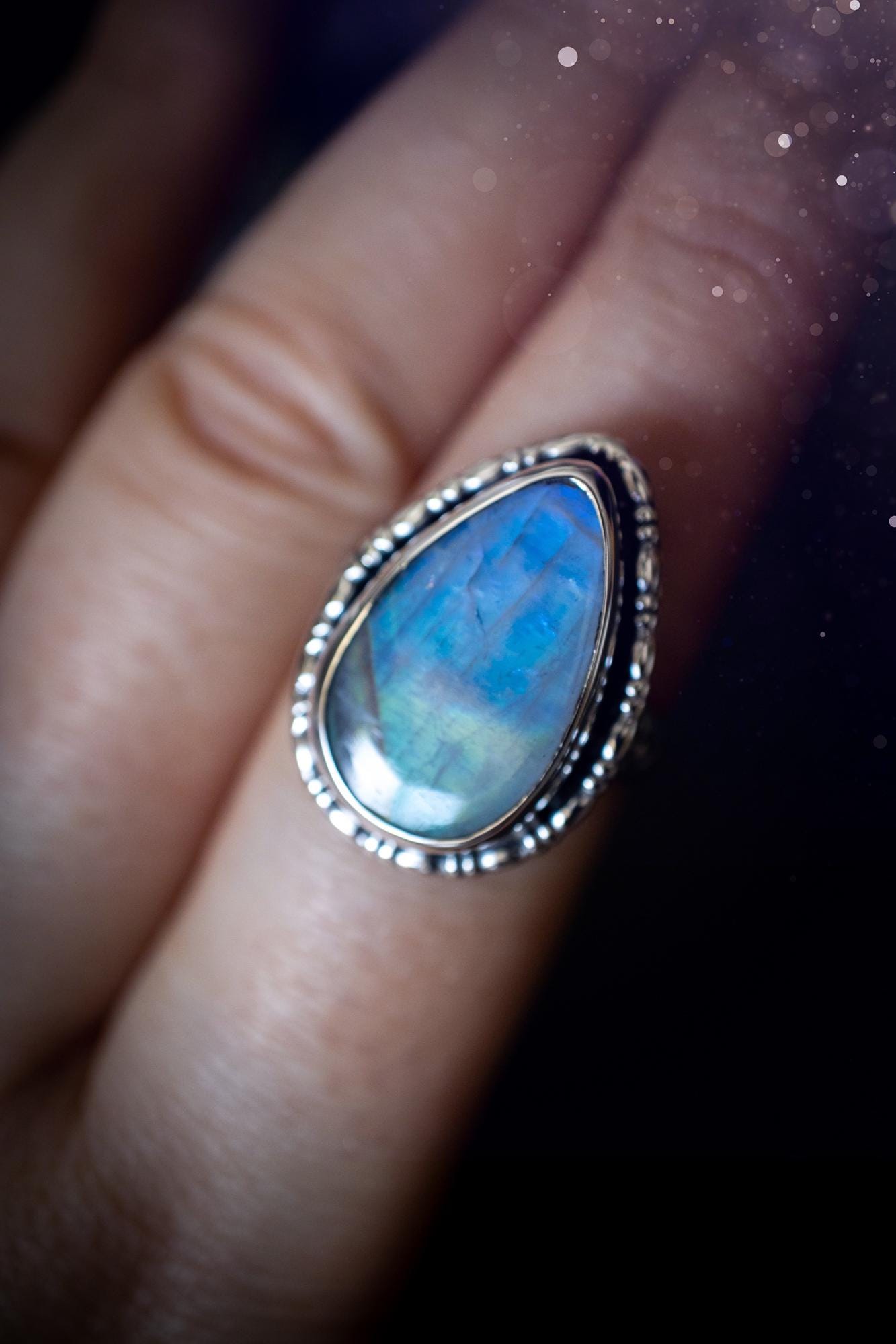A handcrafted rainbow moonstone statement ring with a large, iridescent gemstone glowing with blue and purple flashes. The design blends gothic and whimsical &quot;hoppy&quot; vibes, with bespoke stamping etched into the sterling and fine silver shank. The silver band features intricate detailing, adding texture and character, while the moonstone is bezel-set, enhancing its mystical, ethereal aura. A bold and unique artisan piece with personalized charm. Ideal for crystal and jewellery lovers, h