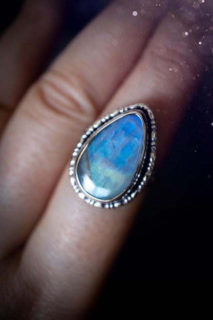 A handcrafted rainbow moonstone statement ring with a large, iridescent gemstone glowing with blue and purple flashes. The design blends gothic and whimsical &quot;hoppy&quot; vibes, with bespoke stamping etched into the sterling and fine silver shank. The silver band features intricate detailing, adding texture and character, while the moonstone is bezel-set, enhancing its mystical, ethereal aura. A bold and unique artisan piece with personalized charm. Ideal for crystal and jewellery lovers, h