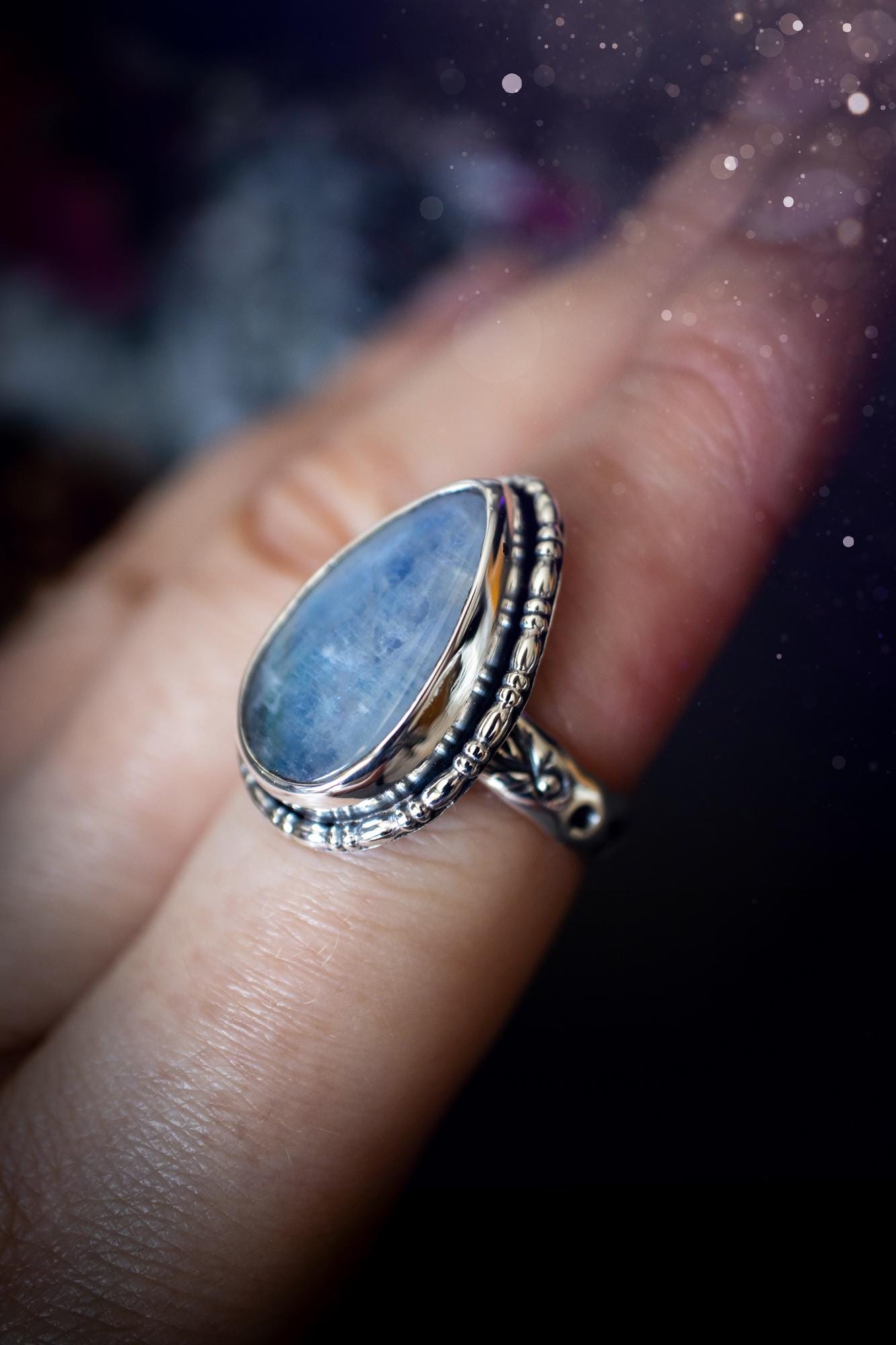 A handcrafted rainbow moonstone statement ring with a large, iridescent gemstone glowing with blue and purple flashes. The design blends gothic and whimsical &quot;hoppy&quot; vibes, with bespoke stamping etched into the sterling and fine silver shank. The silver band features intricate detailing, adding texture and character, while the moonstone is bezel-set, enhancing its mystical, ethereal aura. A bold and unique artisan piece with personalized charm. Ideal for crystal and jewellery lovers, h