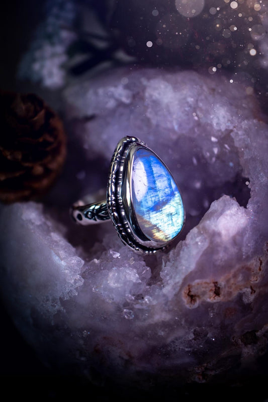 A handcrafted rainbow moonstone statement ring with a large, iridescent gemstone glowing with blue and purple flashes. The design blends gothic and whimsical &quot;hoppy&quot; vibes, with bespoke stamping etched into the sterling and fine silver shank. The silver band features intricate detailing, adding texture and character, while the moonstone is bezel-set, enhancing its mystical, ethereal aura. A bold and unique artisan piece with personalized charm. Ideal for crystal and jewellery lovers, h