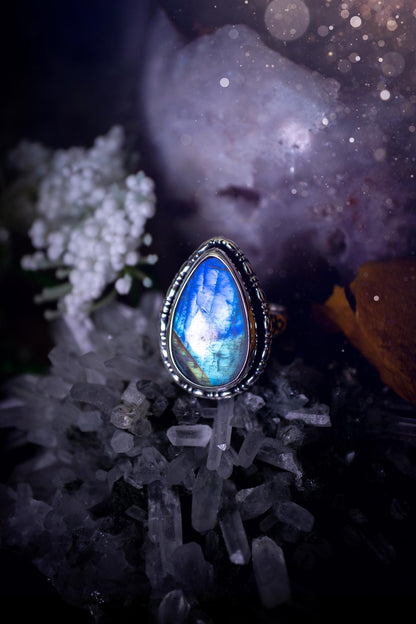 A handcrafted rainbow moonstone statement ring with a large, iridescent gemstone glowing with blue and purple flashes. The design blends gothic and whimsical &quot;hoppy&quot; vibes, with bespoke stamping etched into the sterling and fine silver shank. The silver band features intricate detailing, adding texture and character, while the moonstone is bezel-set, enhancing its mystical, ethereal aura. A bold and unique artisan piece with personalized charm. Ideal for crystal and jewellery lovers, h