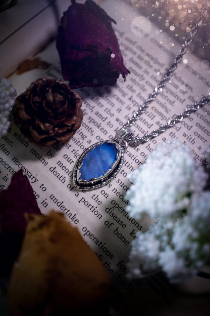 A handmade rainbow moonstone pendant necklace featuring a glowing, iridescent moonstone with blue and purple flashes. The stone is set in a scallop bezel, framed by bespoke gothic gallery wire that adds both hippy and witchy vibes. Crafted from sterling and fine silver, the pendant hangs on a silver chain, exuding mystical charm and alternative style. A one-of-a-kind piece with gothic, and magical energy. The ideal gift for crystal and jewellery lovers. Moonstone is the birthstone for June.