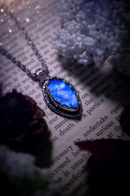 A handmade rainbow moonstone pendant necklace featuring a glowing, iridescent moonstone with blue and purple flashes. The stone is set in a scallop bezel, framed by bespoke gothic gallery wire that adds both hippy and witchy vibes. Crafted from sterling and fine silver, the pendant hangs on a silver chain, exuding mystical charm and alternative style. A one-of-a-kind piece with gothic, and magical energy. The ideal gift for crystal and jewellery lovers. Moonstone is the birthstone for June.