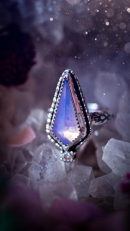 Handmade lavender rose quartz ring crafted from sterling silver and fine silver. The soft lavender gemstone is set within a decorative serrated bezel and surrounded by intricate outer gallery wire detailing. A sparkling aurora cubic zirconia accent adds flashes of rainbow colour. Inspired by fairy and woodland aesthetics, this one-of-a-kind artisan ring features a beautifully handcrafted design and is a UK size O.