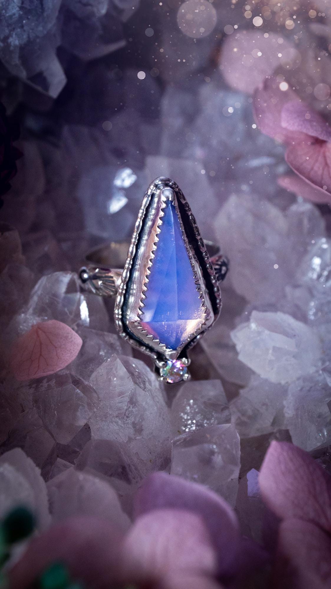 Handmade lavender rose quartz ring crafted from sterling silver and fine silver. The soft lavender gemstone is set within a decorative serrated bezel and surrounded by intricate outer gallery wire detailing. A sparkling aurora cubic zirconia accent adds flashes of rainbow colour. Inspired by fairy and woodland aesthetics, this one-of-a-kind artisan ring features a beautifully handcrafted design and is a UK size O.