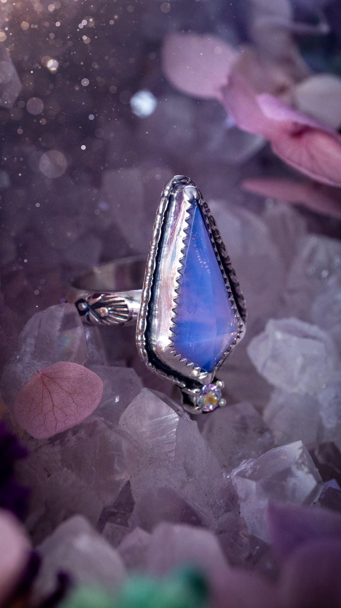 Handmade lavender rose quartz ring crafted from sterling silver and fine silver. The soft lavender gemstone is set within a decorative serrated bezel and surrounded by intricate outer gallery wire detailing. A sparkling aurora cubic zirconia accent adds flashes of rainbow colour. Inspired by fairy and woodland aesthetics, this one-of-a-kind artisan ring features a beautifully handcrafted design and is a UK size O.