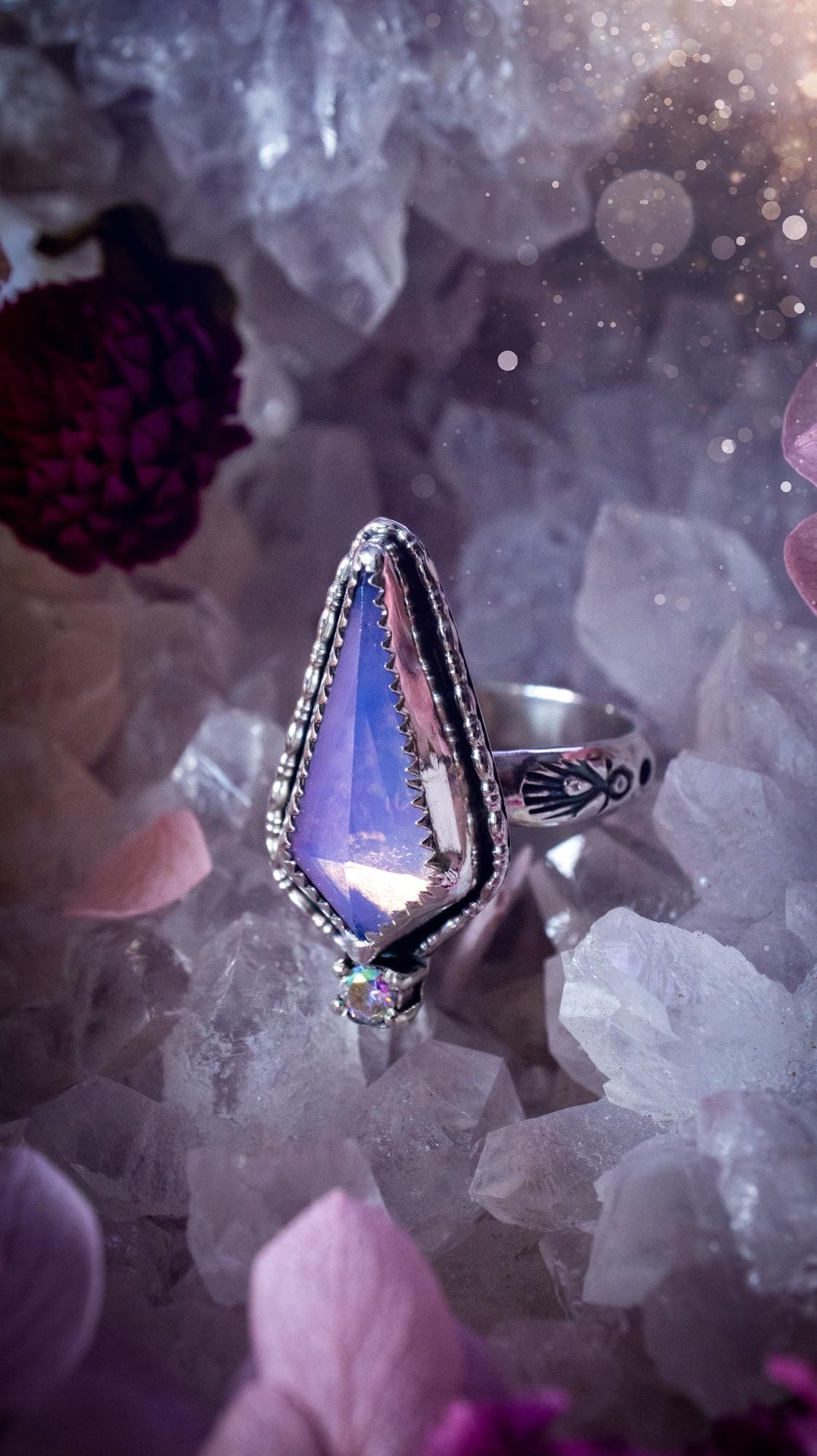 Handmade lavender rose quartz ring crafted from sterling silver and fine silver. The soft lavender gemstone is set within a decorative serrated bezel and surrounded by intricate outer gallery wire detailing. A sparkling aurora cubic zirconia accent adds flashes of rainbow colour. Inspired by fairy and woodland aesthetics, this one-of-a-kind artisan ring features a beautifully handcrafted design and is a UK size O.