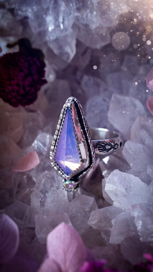 Handmade lavender rose quartz ring crafted from sterling silver and fine silver. The soft lavender gemstone is set within a decorative serrated bezel and surrounded by intricate outer gallery wire detailing. A sparkling aurora cubic zirconia accent adds flashes of rainbow colour. Inspired by fairy and woodland aesthetics, this one-of-a-kind artisan ring features a beautifully handcrafted design and is a UK size O.
