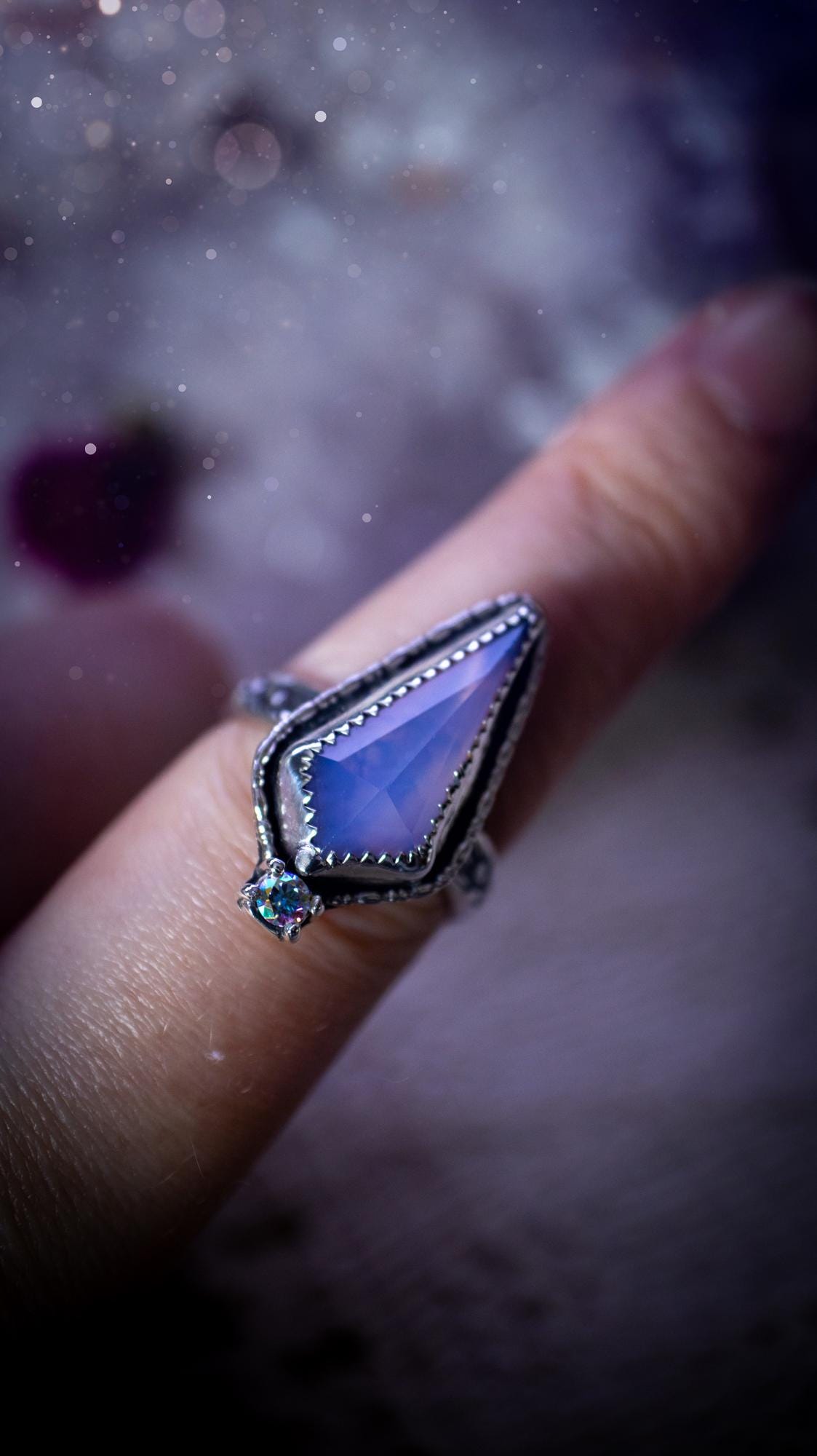 Handmade lavender rose quartz ring crafted from sterling silver and fine silver. The soft lavender gemstone is set within a decorative serrated bezel and surrounded by intricate outer gallery wire detailing. A sparkling aurora cubic zirconia accent adds flashes of rainbow colour. Inspired by fairy and woodland aesthetics, this one-of-a-kind artisan ring features a beautifully handcrafted design and is a UK size O.